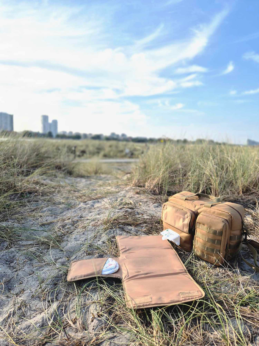 Pusletaske fra Scandinavianbabygear i farven 'khaki', stående på Amagerstrand i København. I baggrunden ses natur og himmel.