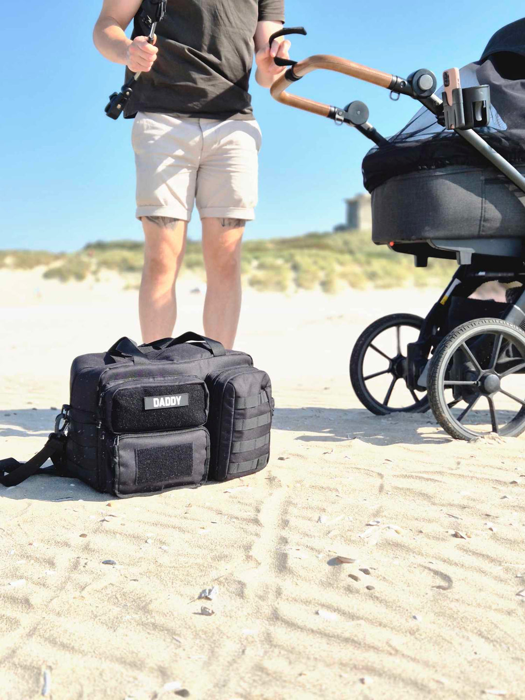 Pusletaske fra Scandinavianbabygear i farven 'sort', stående på en strand i Jylland. I baggrunden ses en far og en barnevogn.