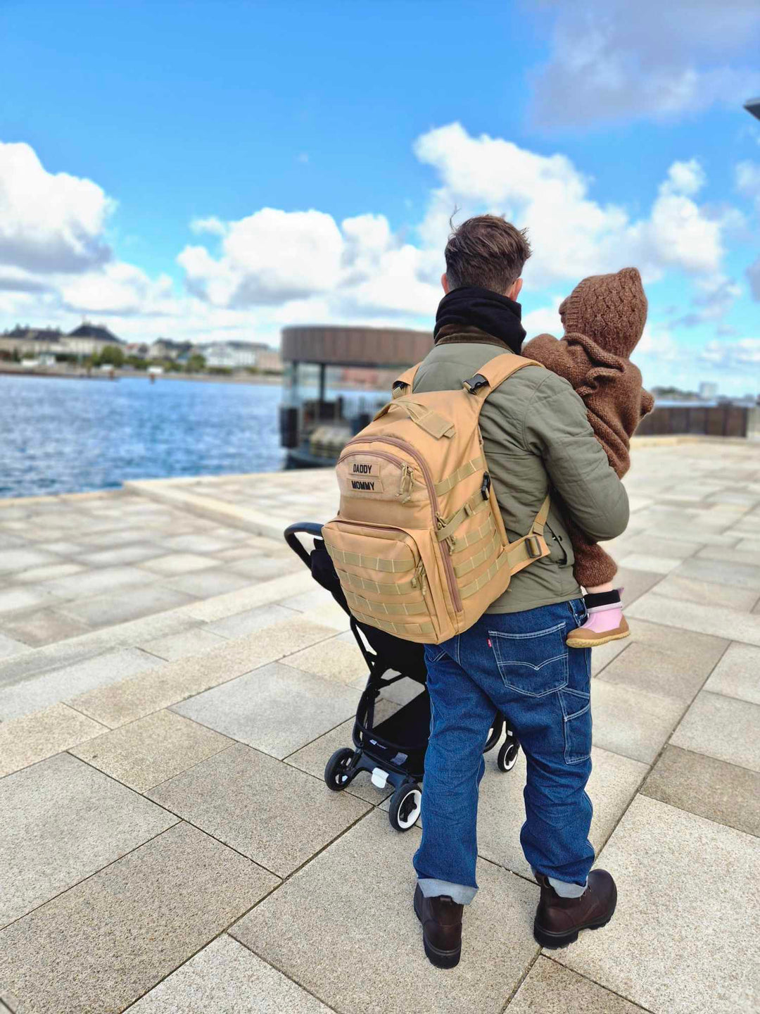 Far med sit barn står ved Operaen i København. Pusletasken er fra Scandinavianbabygear i farven 'khaki'. I baggrunden ses en barnevogn.